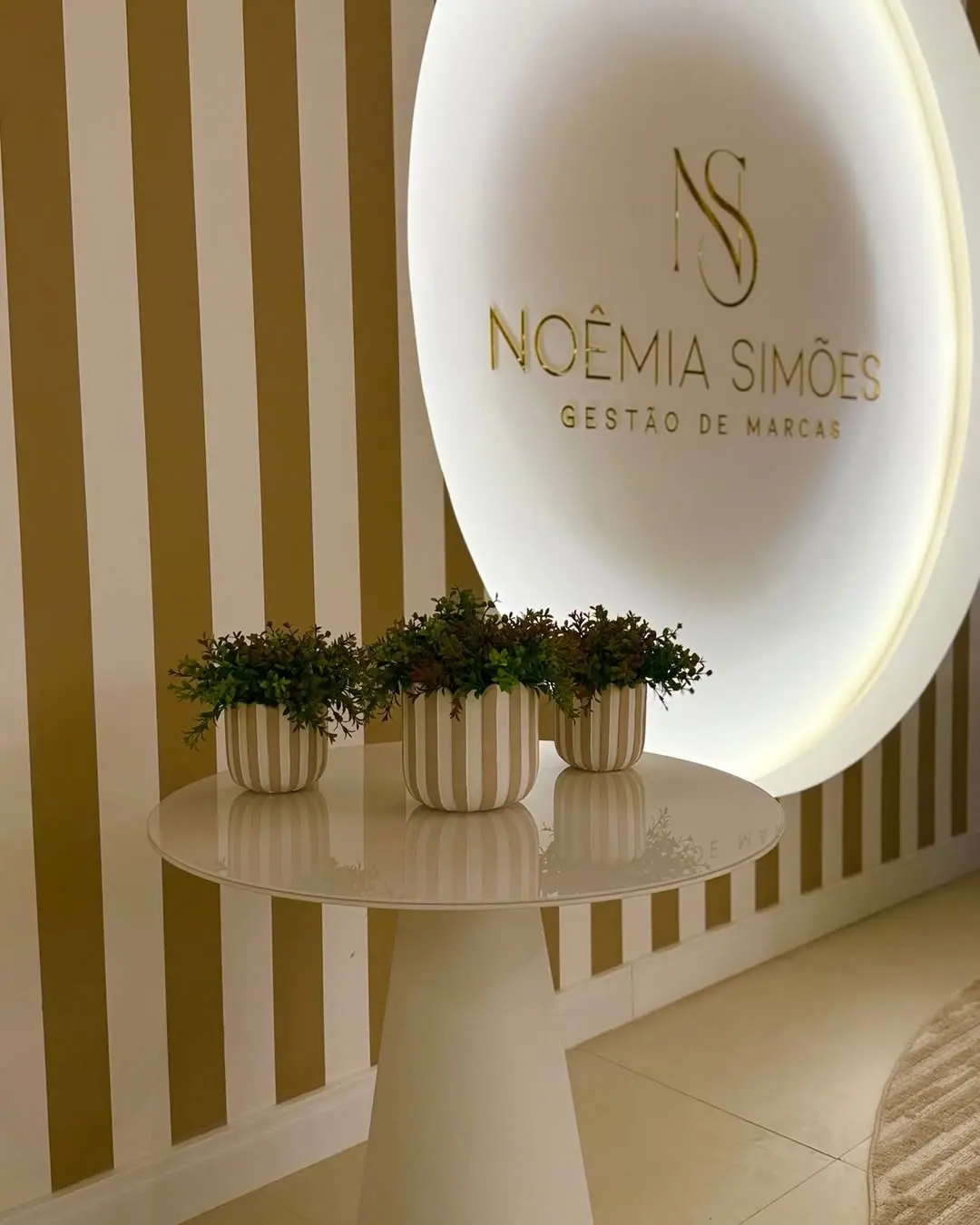 Parede de ripas com o logo Noêmia Simões iluminado e vasos de plantas
